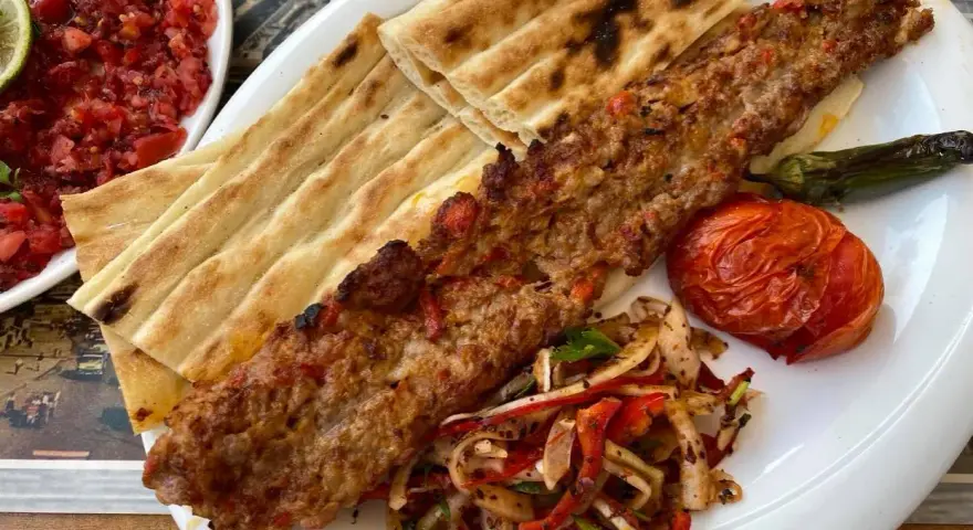 Pyszne tureckie jedzenie: kebab z mięsa mielonego, grillowany pomidor, papryka i świeże pieczywo. Obok sałatka z pomidorów.