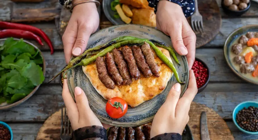 Ręce trzymają talerz z grillowanymi **ćevapi** na płaskim chlebie, ozdobiony papryczkami i pomidorem. Obok sałatka i inne dania.